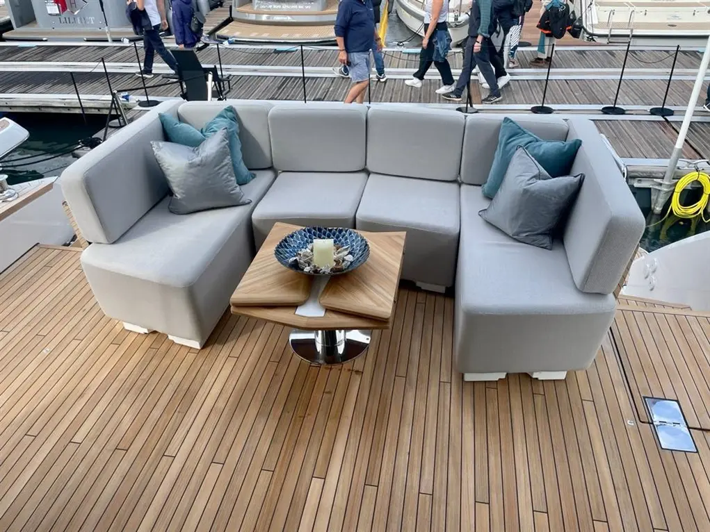 Modern boat seating area with grey cushions, a wooden table, and a decorative bowl, surrounded by a wooden deck and people.